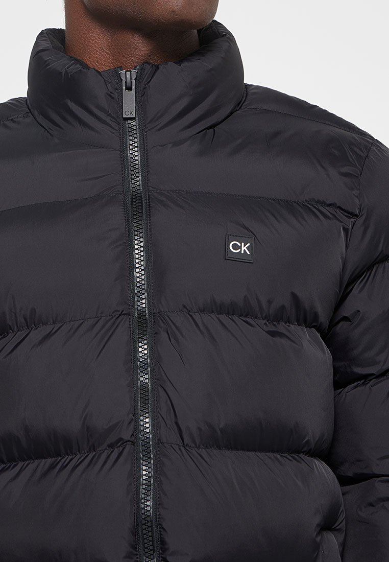 Schwarze Pufferjacke mit hohem Kragen, gestepptem Design und einem Reißverschluss vorne. Hat ein kleines CK-Logo auf der Brust. Glatte Textur.