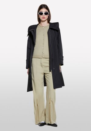 Manteau long noir, cardigan boutoné vert clair, et pantalon large beige avec fermetures éclair dorées. Les accessoires comprennent des lunettes de soleil blanches et des chaussures noires.