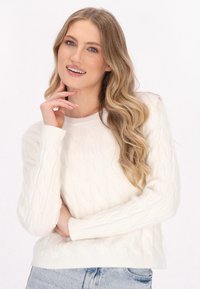 Maglione a trecce di colore crema con scollatura tonda e polsini a coste, abbinato a jeans azzurri chiari. Motivo texturizzato su tutta la parte del capo.