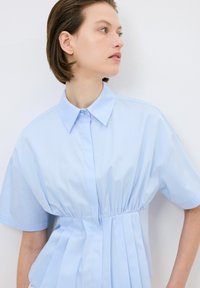 Camicia plissettata azzurro chiaro con maniche corte, colletto button-down e vita arricciata. Realizzata in tessuto liscio con un design strutturato.