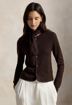 Femme portant un pull marron foncé boutonné avec une écharpe assortie et un pantalon blanc plissé taille haute, mains dans les poches, regardant vers le bas.