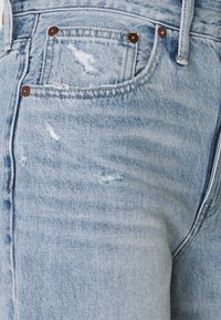 Světle modré džíny z denimu s opotřebovaným vzhledem. Obsahují třepivé části, kontrastní hnědé kovové nýty a klasický pětikapsový design.