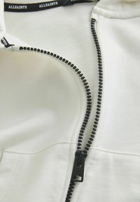 Vêtement blanc à fermeture éclair, avec une texture côtelée, doté d'une fermeture éclair noire contrastante et d'une languette de tirage. Comprend un détail de couture de poche visible.