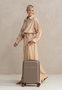 Een lachende vrouw in een beige outfit trekt een stijlvolle koffer met gouden accenten, tegen een neutrale achtergrond.