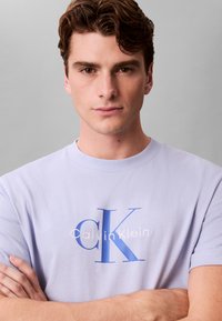 Ung mand med korslagte arme iført en lyse lilla Calvin Klein t-shirt mod en ensfarvet grå baggrund.