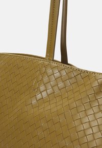 Spleciona torba na ramię w musztardowej skórze, charakteryzująca się gładką teksturą i podwójnymi uchwytami. Projekt zawiera geometryczny wzór.