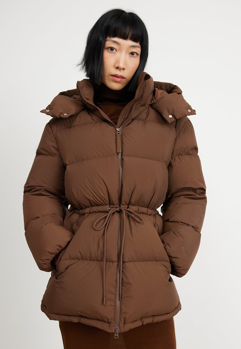 Brązowa kurtka puchowa z wcięciem w talii, zamkiem błyskawicznym i wysokim kołnierzem. Posiada pikowany wzór oraz napy ozdobne na kapturze.