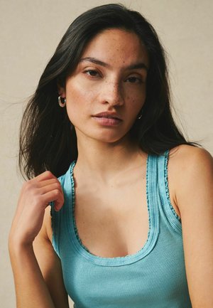 Ung kvinde med langt mørkt hår og fregner iført en turkis ribbet tanktop, poserer med den ene hånd nær sin skulder mod en beige baggrund.