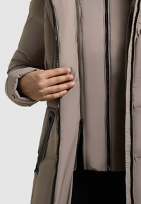 Chaqueta acolchada de color marrón claro con una superficie lisa, que cuenta con dobles cremalleras frontales y un forro interior texturizado. Acentos de hardware negro visibles.