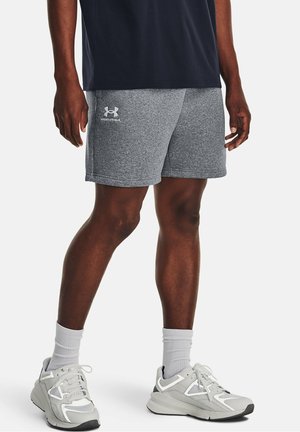 Mand iført grå Under Armour shorts, hvide crew sokker og hvide og grå atletiksko, stående mod en hvid baggrund.