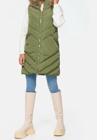 Olijfgroene mouwloze puffervest met een capuchon, voorzien van een chevronpatroon, zijzakken en goudkleurige ritsen, gecombineerd met blauwe jeans en beige laarzen.
