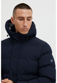 Tmavě modrá prošívaná bunda s vysokým límcem, černými patentními knoflíky a logem na rukávu. Hladká látka, prošívaný vzor.