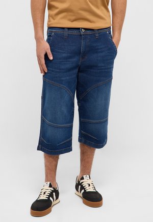 Man wearing knee-length dark blue denim shorts, black and white sneakers, and a brown t-shirt, standing with hands in pockets.