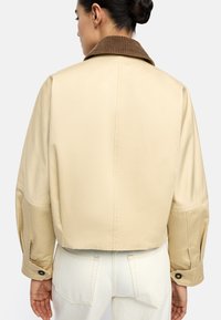 Kobieta widziana z tyłu, ubrana w beżową kurtkę typu cropped z brązowym kołnierzem z kordurowej tkaniny oraz w off-white wysokiej talii spodnie.