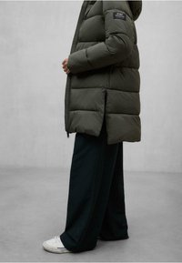 Persona vestida con una chaqueta acolchada verde oscuro con capucha, pantalones anchos negros y zapatillas blancas, de pie contra un fondo gris liso.