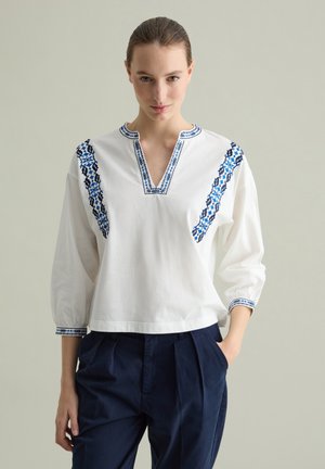 White cotton blouse with a V-neckline, featuring blue and black floral embroidery along the shoulders and sleeves, textured cuffs, and a loose fit.