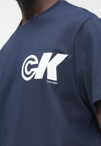 Dark-skinned person wearing a navy blue t-shirt with large white "CK" Calvin Klein logo on the chest, cropped at neck and shoulder.