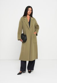Cappotto lungo verde oliva con rever a scollar, maniche ampie e tasca anteriore. Abbinato a pantaloni navy a gamba larga e una borsa a mano nera.