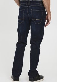 Jeans de mezclilla de un oscuro azul, con un corte ajustado, que presentan dos bolsillos traseros y costuras en contraste. Llevados con zapatillas negras.