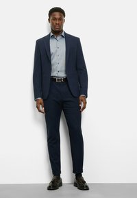 Navy suit jacket over a light blue button-down shirt, paired with dark slim-fit trousers and black dress shoes. Smooth textures throughout.