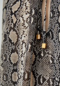 Gros plan sur un tissu à motif peau de serpent avec deux cordons noirs suspendus se terminant par de petites perles cubes dorées.