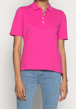 Femme portant un polo rose vif à manches courtes avec des boutons et un jean bleu clair, debout devant un fond uni.