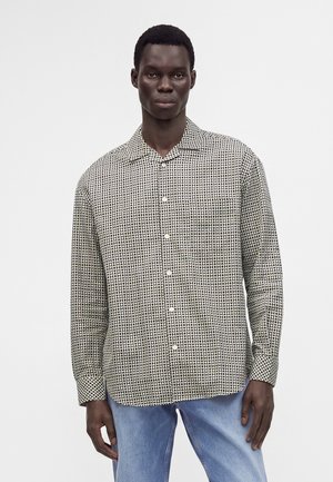 Homme portant une chemise boutonnée à manches longues avec un motif géométrique noir et blanc et un jean bleu clair, debout devant un fond uni.