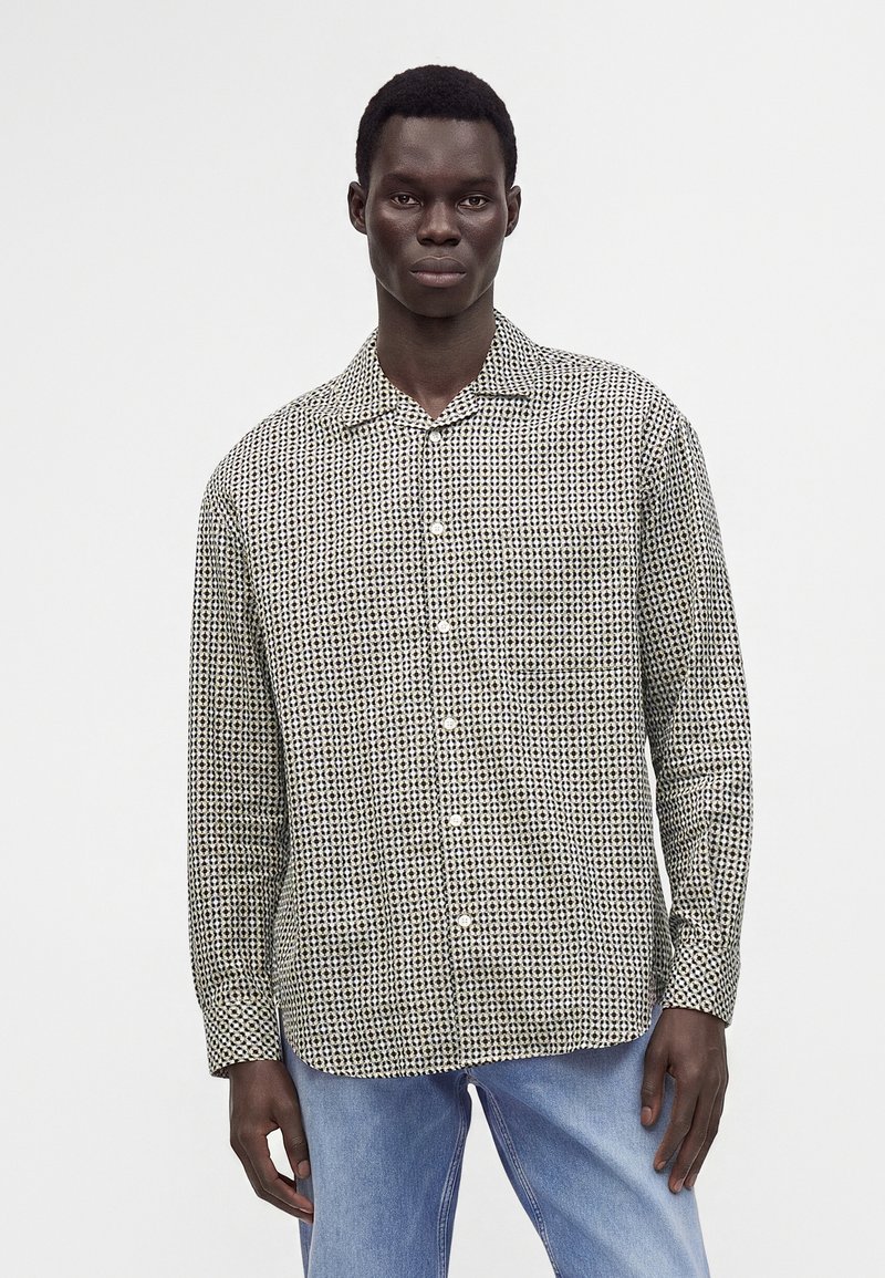 Homme portant une chemise boutonnée à manches longues avec un motif géométrique noir et blanc et un jean bleu clair, debout devant un fond uni.