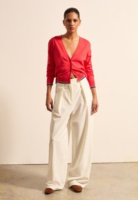 Rote kurze Strickjacke mit V-Ausschnitt, die über einen Knopfverschluss und marineblaue Akzente verfügt. Kombiniert mit weit geschnittenen weißen Hosen und braunen Schuhen.