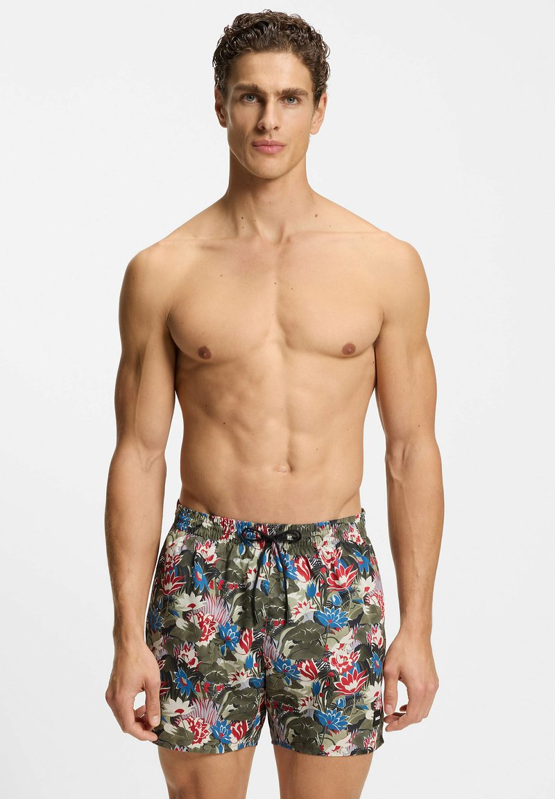 Uomo senza maglietta con capelli ricci che indossa pantaloncini da bagno a motivo floreale nei colori rosso, blu e verde, in piedi contro uno sfondo semplice.