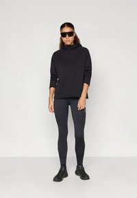 Žena s kudrnatými vlasy, oblečená v černé oversized mikině, černých legínách, černých teniskách a velkých černých slunečních brýlích, stojící proti jednobarevnému pozadí.