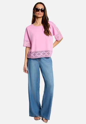 Femme portant un haut rose à manches courtes avec des détails en dentelle et un jean bleu à jambes larges, accessoirisée avec des lunettes de soleil foncées et des boucles d'oreilles.