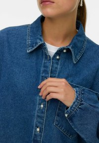 Camicia di jeans blu, con colletto a punta, bottoni simili a perle e taschino sul petto. Tessuto a trama visibile e polsini delle maniche arricciati.