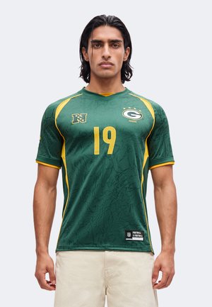 Homme aux cheveux noirs mi-longs, portant un maillot vert et jaune des Green Bay Packers avec le numéro 19, debout devant un fond uni.