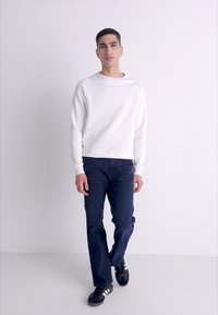 Sudadera blanca con cuello redondo, jeans azules y zapatillas negras. Tela suave con un ajuste casual, de pie sobre un fondo gris claro.