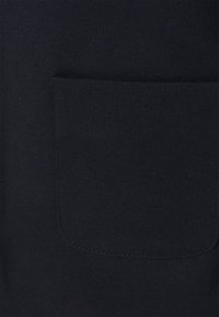 Gros plan sur un tissu noir montrant une poche plaquée rectangulaire unie avec des coins inférieurs arrondis et des coutures visibles.