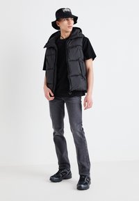 Gilet matelassé noir avec capuche sur un t-shirt noir, associé à un jean gris et des baskets noires. Le chapeau seau arbore un logo blanc.