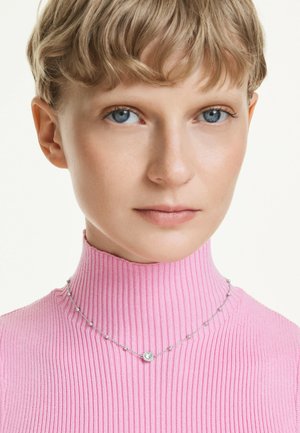 Primer plano de una persona con cabello corto y rubio que lleva un cuello alto rosa acanalado y un delicado collar de plata con pequeñas cuentas y un colgante circular.