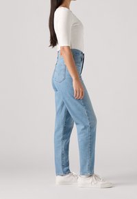 Femme debout portant un jean taille haute bleu clair, un haut blanc côtelé avec manches aux coudes et des baskets blanches sur un fond clair uni.