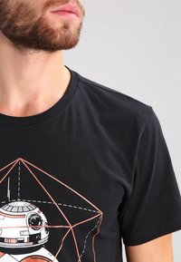 Camiseta de algodón negra con un diseño geométrico que presenta una ilustración de un droide en líneas blancas y naranjas. Con mangas cortas y cuello redondo.