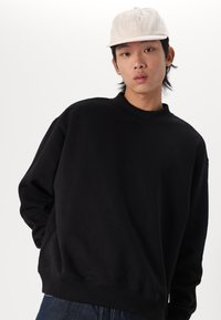 Schwarzer Hoodie mit hohem Kragen, oversized Passform und gerippten Bündchen, kombiniert mit einer hellbeigen Kappe mit gestickten Logo-Details.