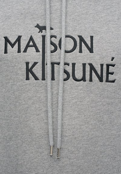 Hanorac gri cu text brodat negru care citește „MAISON KITSUNÉ.” Are șnururi cu vârfuri metalice și o textură de țesătură netedă.