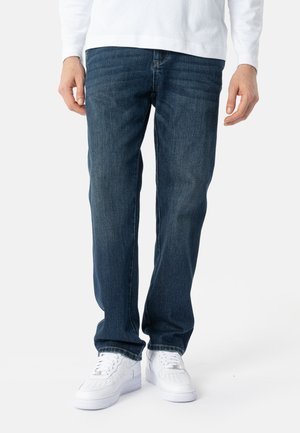 Mann, der dunkelblaue Jeans mit geradem Bein, weißes Langarmshirt und weiße Turnschuhe trägt, steht vor einem schlichten Hintergrund.