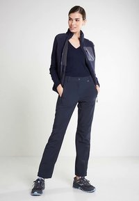 Γυναίκα στέκεται με τα χέρια στις τσέπες, φορώντας ναυτικό μπλε φλις μπουφάν, αντίστοιχο V-neck τοπ, παντελόνι και μαύρα αθλητικά παπούτσια.