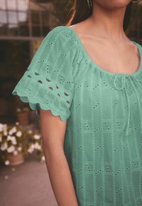 Teal bluse med korte, bølgede ærmer med hulmønster og firkantet halsudskæring. Bånddetalje foran, letvægtsstof.