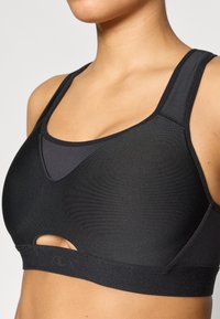 Μαύρο αθλητικό σουτιέν με λείο ύφασμα, φαρδιές τιράντες, λεπτομέρεια με άνοιγμα και καμπυλωτή γραμμή στο ντεκολτέ. Διαθέτει λογότυπο στη ζώνη.