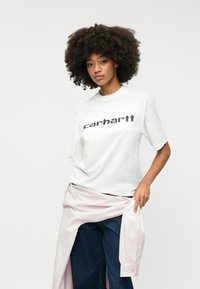 Fehér pamut póló fekete "carhartt" logóval a mellkason, sötét farmernadrággal és világos rózsaszín inggel a derékon megkötve.