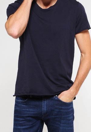 Persoon die een effen donkerblauw t-shirt en blauwe spijkerbroek draagt, met één hand in de zak en de andere die hun nek aanraakt.