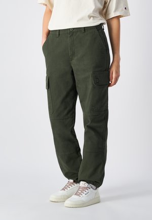 Cargo-Hosen aus dunkelgrünem Baumwollmischgewebe mit Seitentaschen, lockerer Passform und elastischen Bündchen. Kombiniert mit weißen Sneakers für einen Kontrast.