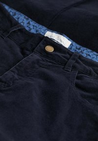 Marina kordflosshosen med en strukturerad yta, med ett mönstrat blått midjeband och en detaljerad metallknapp med varumärke.